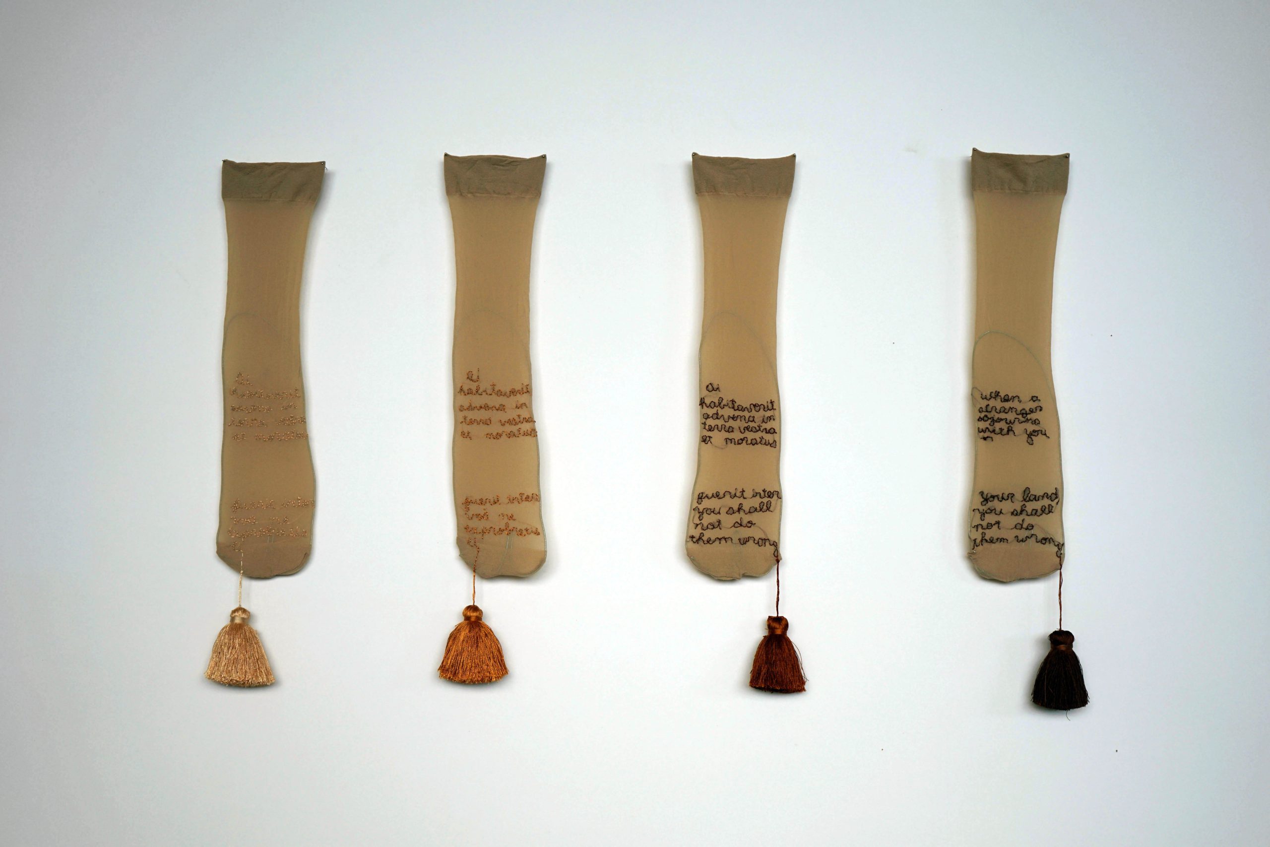 Dirt, Skin and Gold, 2019,Secondhand socks, silk thread embroidery, silk pom poms, 8 x 20 cm each. Socks embroidered with Old Testament text related to rights to land, translated in different ways. 